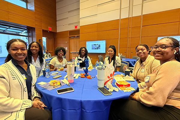 Eight people seated around a round table with a blue tablecloth in a conference room, with food, drinks, and phones on the table and a presentation screen visible in the background.