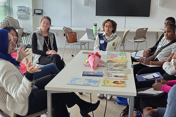 Several people seated around a rectangular table in a classroom, engaged in discussion, with notebooks, illustrated books, and a plush anatomical model placed on the table.