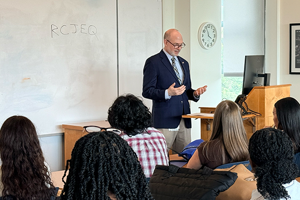 A person standing at a podium speaking to an audience seated in a classroom, with a whiteboard behind them displaying handwritten text and a wall clock nearby.