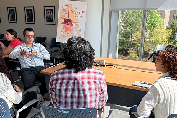 Small group discussion in a classroom, with one person gesturing and a digestive system poster near a window.