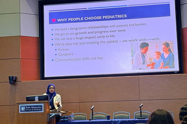 Speaker at a podium presenting a slide titled ‘Why People Choose Pediatrics’ in an auditorium.