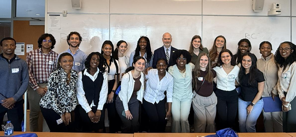 Group of people posing for a photo in a classroom with desks and blue drawstring bags.